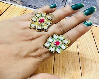 May include: Two gold-toned rings with a floral design. Each ring features a central red stone surrounded by white and green accents. The rings are displayed on a hand with green nail polish.