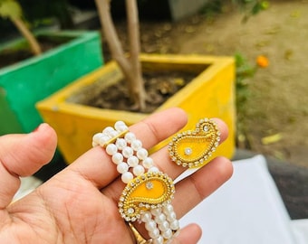 May include: A gold-toned bracelet and matching earrings set. The bracelet features multiple strands of white pearls with a yellow paisley-shaped centerpiece. The earrings have a similar design. The jewelry is embellished with small, clear gemstones.