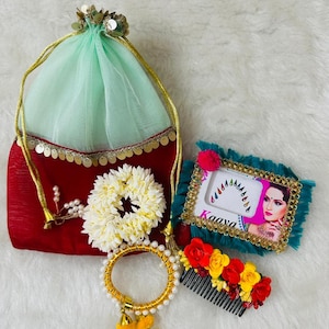 May include: An assortment of Indian wedding accessories, including a red and green pouch, a white floral garland, a gold bangle with pearls, a comb with red and yellow flowers, and a framed image with the word "Kaaya".