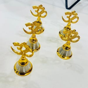 May include: Four gold-colored Om symbols on silver and gold bases. The Om symbols are intricately designed and stand upright. The bases have a bell-like shape. The items are on a white surface.