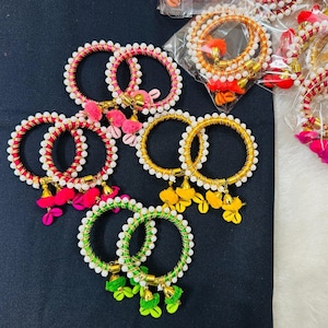 May include: An assortment of colorful bangles with intricate designs. The bangles feature a variety of colors, including red, pink, green, and gold, adorned with white beads and decorative tassels. Some are packaged in clear plastic.