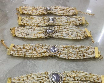 May include: Five beaded bracelets with gold-colored clasps and centerpieces. The bracelets feature off-white and gold beads, with a large, clear, gemstone-like centerpiece. The clasps and centerpieces have a gold finish.