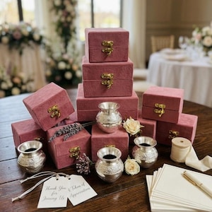 Puede incluir: Cajas de terciopelo rosa empolvado con cierres dorados, jarrones decorativos plateados, rosas blancas y una etiqueta con la frase "Thank You for Joining Us". Dispuestas sobre una mesa de madera oscura.