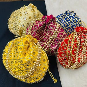 May include: An assortment of five decorative drawstring pouches in various colors: gold, yellow, magenta, blue, and red. Each pouch features a geometric pattern with mirrored accents and is adorned with a beaded trim. The pouches are likely used for carrying small items.