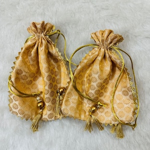 Könnte beinhalten: Zwei goldene Kordelzugbeutel mit floralen Mustern. Jeder Beutel hat eine goldene Quaste und Perlenakzente. Die Kanten sind mit einer dekorativen Goldborte versehen. Die Beutel liegen auf einer weißen, flauschigen Oberfläche.