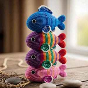 Puede incluir: Una pila de cuatro juguetes de peces hechos a mano con ganchillo. El pez superior es azul, los dos del medio están rayados con varios colores y el pez inferior es rosa. Cada pez tiene una aleta con ribete blanco y un ojo negro. Los juguetes están apilados sobre una superficie de madera.