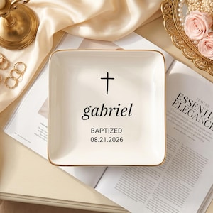 May include: A square white ceramic trinket dish with a gold-toned rim. The center features a black cross icon above the name "gabriel" in a serif font, followed by the text "BAPTIZED" and the date "08.21.2026" printed in black ink.