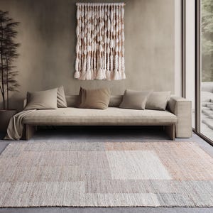 Puede incluir: Una escena de salón de tonos neutros con un sofá beige con varios cojines, un tapiz de pared tejido y una alfombra estampada. La alfombra tiene un diseño geométrico con tonos beige, gris y melocotón. Una planta alta en maceta y una ventana grande completan la habitación.