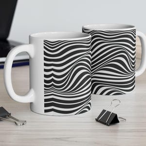 Peut inclure: Deux tasses en céramique blanches avec un motif à rayures ondulées noires et blanches. Les tasses ont une anse standard et sont posées sur une surface en bois de couleur claire. Un ordinateur portable, un crayon et des pinces à reliure sont également visibles.