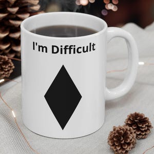 May include: White ceramic mug with the text "I'm Difficult" above a black diamond shape. The mug is filled with a dark liquid and has a white handle. The mug is on a light grey surface with decorative items.