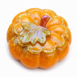 May include: A glossy orange pumpkin-shaped object with a smooth, rounded surface and deep vertical ridges. It features a central iridescent green leaf and a small stem, with embedded gold glitter accents throughout the translucent resin material.