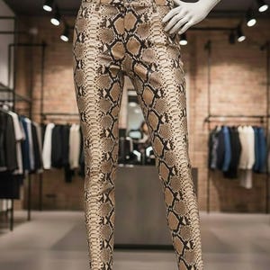以下が含まれることがあります： 茶色と黒の色合いのスネークスキン柄のスリムフィットパンツ。衣料品店内のマネキンに展示されており、詳細なプリントとフィット感をアピールしています。ボタンとジッパーで開閉します。