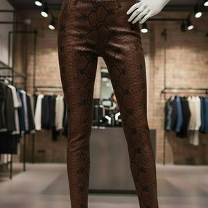 May include: Brown faux leather pants with a snakeskin pattern. The pants are displayed on a mannequin in a clothing store. The pants have a classic five-pocket design and a button and zipper closure.
