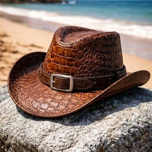 Könnte beinhalten: Ein brauner Cowboyhut mit einem strukturierten, krokodilartigen Muster liegt auf einem Felsen in der Nähe des Strandes. Der Hut hat ein Lederband mit einer silbernen Schnalle. Der Hintergrund zeigt das Meer und einen Sandstrand.