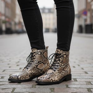 Puede incluir: Botines con estampado de piel de serpiente en tonos marrón, beige y negro. Las botas tienen cordones negros y un tacón bajo negro. La persona lleva vaqueros negros. Calzado elegante.