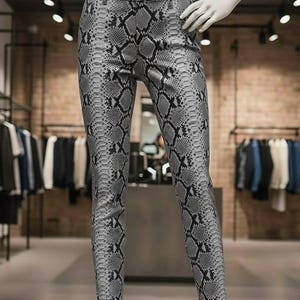 Op de afbeelding: Grijze en zwarte broek met slangenprint. De broek wordt getoond op een mannequin in een kledingwinkel. De broek heeft een klassiek ontwerp met vijf zakken en een getailleerde pasvorm. Het slangenpatroon is gedetailleerd en realistisch.