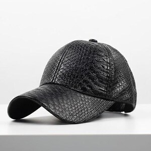 May include: A black baseball cap with a textured, snake-skin pattern. The cap has a curved brim and a rounded crown. The hat is made of a dark material and sits on a white surface.