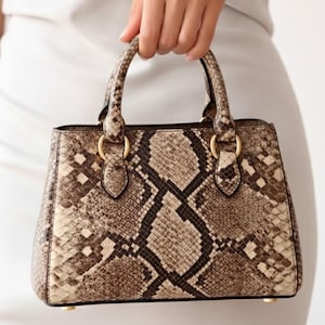 Op de afbeelding: Handtas met slangenprint en een gestructureerde vorm. De tas heeft een handvat aan de bovenkant, goudkleurige ringen en een rechthoekig silhouet. Het kleurenpalet omvat bruin, beige en zwart.