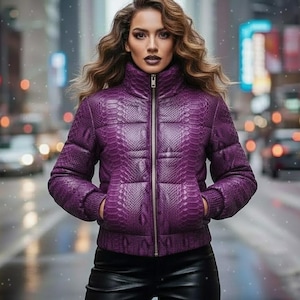 May include: A quilted, purple puffer jacket with a snakeskin pattern. The jacket has a high collar and a silver zipper. The person is wearing black leather pants. The background is a city street with blurred lights.