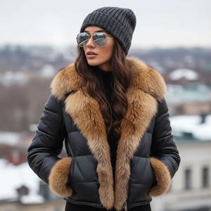 Puede incluir: Chaqueta acolchada negra con cuello y puños de piel marrón. La persona de la imagen lleva un gorro de punto gris y gafas de sol de aviador. La chaqueta tiene cierre de cremallera.