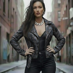 May include: A woman wearing a snakeskin-patterned jacket, black top, and black jeans. The jacket has a notched collar and a belt. A silver chain necklace is worn around the neck. The background is a blurred city street.