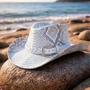 以下が含まれることがあります： 白いカウボーイハット、ヘビ革模様。大きな岩の上に置かれています。白い革バンド、シルバーバックル、編み込みディテール付き。背景にはビーチと海が写っています。