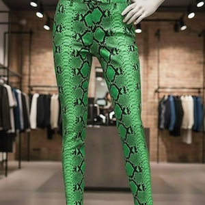 Könnte beinhalten: Grüne Hose mit Schlangenmuster an einer Schaufensterpuppe. Die Hose hat eine leuchtend grüne Farbe mit einem schwarzen Schuppenmuster. Die Hose wird in einem Bekleidungsgeschäft präsentiert, mit Kleiderständern im Hintergrund.
