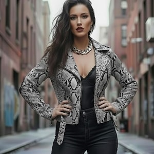 May include: A snakeskin-patterned jacket in shades of gray and white, featuring a classic biker style with a notched collar and asymmetrical zipper. The jacket is paired with a black top, jeans, and a chunky gold chain necklace.