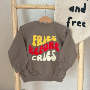 May include: A brown long-sleeve sweatshirt with a crew neckline and ribbed cuffs and hem. The back features the text "FRIES BEFORE CRIES" printed in a wavy, bold font with "FRIES" in yellow, "BEFORE" in red, and "CRIES" in cream.