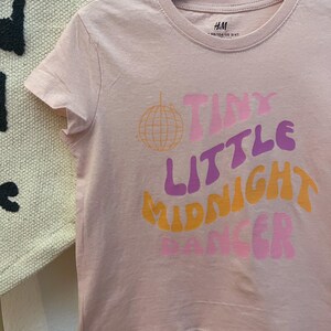 以下が含まれることがあります： レトロなフォントで「Tiny Little Midnight Dancer」の文字が入った薄いピンク色のTシャツ。ディスコボールのグラフィックも特徴です。木製のハンガーにかかっています。