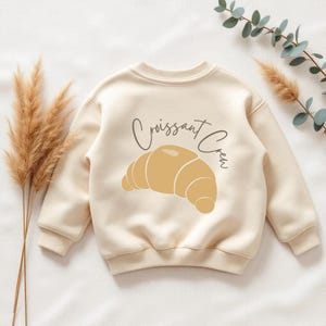 Op de afbeelding: Crèmekleurige sweatshirt met een afbeelding van een croissant en de tekst "Croissant Crew" in een scriptlettertype. Het ontwerp is in tinten geel en bruin. De sweatshirt heeft een ronde hals en lange mouwen.