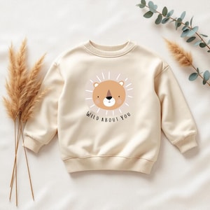 Peut inclure: Sweat-shirt crème avec un motif de lion et le texte "WILD ABOUT YOU". Le lion a un visage marron, un nez noir et une crinière beige clair. Le sweat-shirt a un col rond et des poignets et un ourlet côtelés.