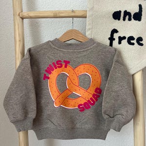 May include: A heathered brown long-sleeve sweatshirt featuring a large orange pretzel graphic on the front. The text "TWIST SQUAD" is printed in bold pink letters curving around the pretzel. The garment has a ribbed crew neckline and cuffs.