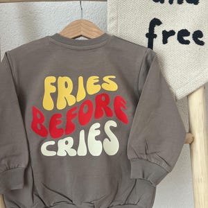 Könnte beinhalten: Ein braunes Sweatshirt mit dem Text "FRIES BEFORE CRIES" in Gelb, Rot und Weiß. Das Sweatshirt hängt an einem Holzbügel. Im Hintergrund ist ein Banner mit den Worten "and free" zu sehen.