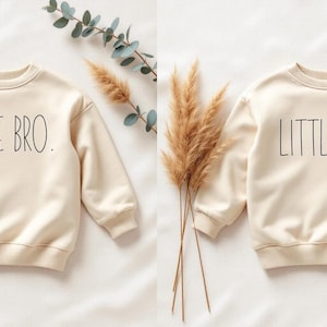 Könnte beinhalten: Zwei cremefarbene Sweatshirts, eines mit "LITTLE BRO." und das andere mit "LITTLE SIS." in Schwarz bedruckt. Die Sweatshirts sind langärmlig und haben gerippte Bündchen. Dekorative Zweige mit Grün und getrockneten Gräsern sind zwischen den Sweatshirts angeordnet.