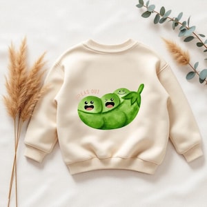 Puede incluir: Sudadera de color crema con una ilustración caprichosa de tres guisantes verdes en una vaina. Los guisantes tienen caras sonrientes y las palabras "PEAS OUT" están impresas encima de la vaina. La sudadera tiene mangas largas y cuello redondo.