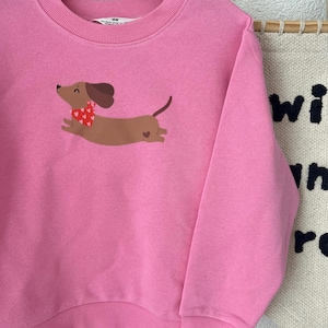 Könnte beinhalten: Ein pinkfarbenes Sweatshirt mit einem Cartoon-Dackel. Der Hund ist braun mit einem roten Schal mit Herzmuster und einem kleinen Herz an der Seite. Das Sweatshirt hängt an einem Holzbügel.