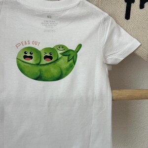 Peut inclure: T-shirt blanc avec un motif de gousse de pois verte et trois petits pois souriants. Le texte "PEAS OUT" est imprimé au-dessus du motif en orange clair. Le t-shirt est suspendu à un cintre en bois.