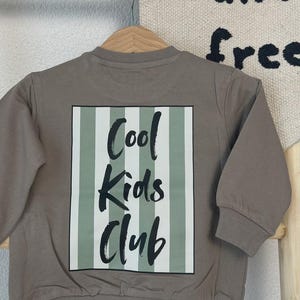 Puede incluir: Una sudadera de color topo con mangas largas, colgada de una percha de madera. La parte trasera de la sudadera presenta un diseño rectangular con rayas verticales verdes y blancas. Las palabras "Cool Kids Club" están impresas en negro.