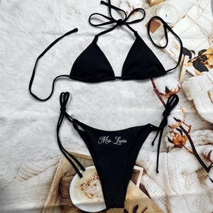 May include: A black bikini set with a triangle top and tie-side bottoms. The bottoms have the embroidered text "Mrs. Levin". The bikini is displayed on a white, textured surface with decorative elements.