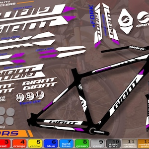 May include: A collection of bicycle stickers in white and purple, including the brand name "GIANT". The set includes stickers for the frame, fork, and other parts, with various sizes and designs. The image also shows a black bicycle frame.