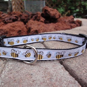 May include: A white dog collar with a black border and a repeating pattern of yellow and black bees. The collar has a silver metal buckle and a black plastic clasp.