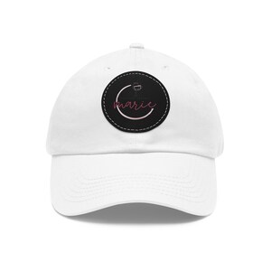 Op de afbeelding: Witte baseballcap met een gebogen klep. Een zwarte ronde patch op de voorkant toont een wijnglasontwerp en de naam "marie" in roze script. De cap is gemaakt van een zachte stof en heeft een verstelbare band.