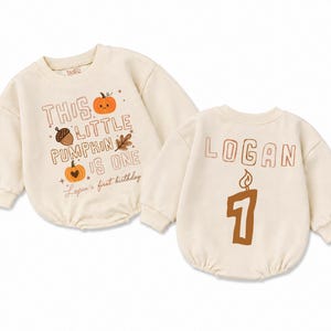 May include: Two cream-colored long-sleeve rompers. The left romper features the text "THIS LITTLE PUMPKIN IS ONE" with pumpkin, acorn, and leaf graphics. The right romper displays the name "LOGAN" above a large brown number one candle graphic.