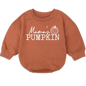 May include: A rust-colored long-sleeve sweatshirt romper with a ribbed crew neckline and cuffs. The front features white text that reads Mama's PUMPKIN, accompanied by a simple white line-art illustration of a pumpkin positioned to the right.