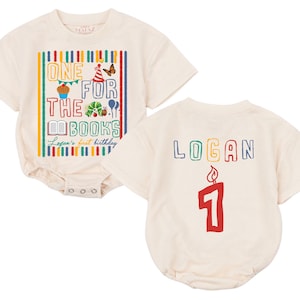 May include: An off-white short-sleeve bodysuit featuring a colorful graphic with the text ONE FOR THE BOOKS and Logan's first birthday. The design includes a cupcake, caterpillar, and book. The back displays the name LOGAN above a red candle number 1.
