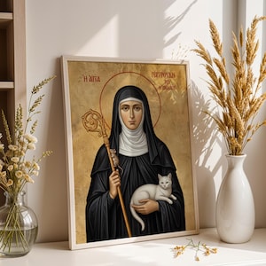 Op de afbeelding: Een religieus icoon in een lijst met een vrouw in een zwarte habijt en witte sluier, die een witte kat en een gouden staf vasthoudt. Het icoon heeft een gouden halo en tekst in een vreemde taal. Het kunstwerk wordt op een wit oppervlak getoond.