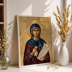 Pode incluir: Ícone religioso emoldurado com uma mulher com um véu azul e uma auréola. O ícone tem texto grego e um fundo dourado. A mulher segura um pergaminho e veste vestes roxas e azuis.