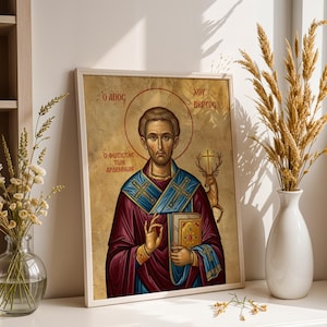 May include: Framed religious icon featuring a saintly figure with a halo, holding a book and a cross. The icon has a gold background and is framed in a white wooden frame. The figure is dressed in a burgundy robe with blue and gold accents. The text in the image is in Greek.