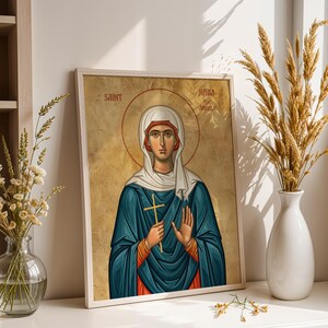 May include: A framed religious icon print featuring Saint Junia with a gold background. The figure wears a white head covering and a blue robe, holding a small gold cross in one hand while the other is raised. The text reads Saint Junia.